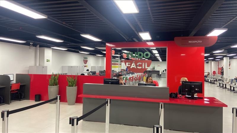 Foto horizontal colorida mostra ambiente interno da unidade do Tudo Fácil no Shopping Passo Fundo. Em primeiro plano um balccão de antedimento vermelho com a logo do Tudo Fácil tem tres atendentes sentados em frente a computadores. Ao fundo estão cadeiras para espera e ghichês de atendimento