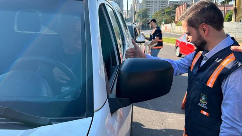 Foto horizontal colorida mostra vice-governador fazendo sinal de legal na janela de um carro branco para motorista sorridente