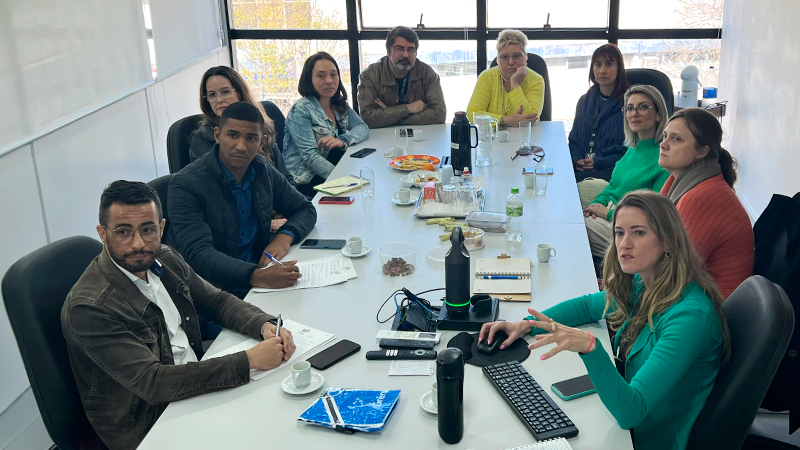 pessoas reunidas em volta de uma mesa retangular com janela ampla ao fundo