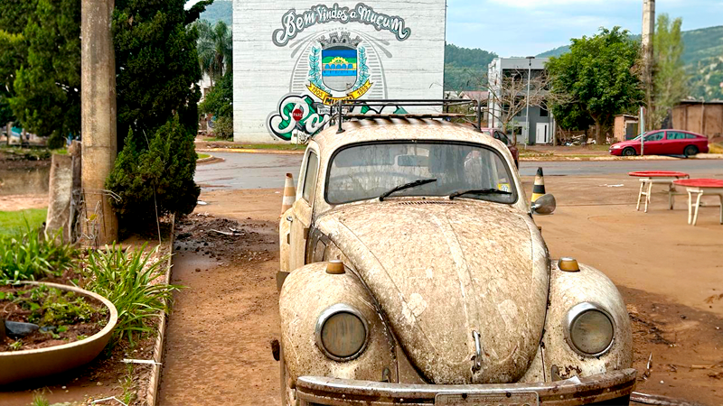 fusca sujo de lama ao centro da imagem, em praça com barro e ao fundo placa onde está escrito bem-vindos a Muçum