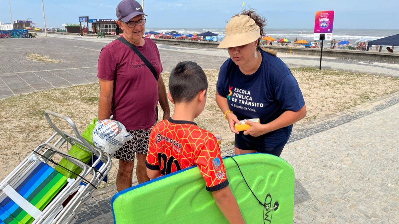 Foto horizontal colorida mostra moça de camiseta e bone conversando com uma criança segurando um bodyboard e um senhor levando cadeiras de praia com o mar ao fundo