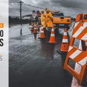 Pista de rodovia interditada por cavalete e cones e agentes de capa de chuva laranja, água em um dos lados da pista.