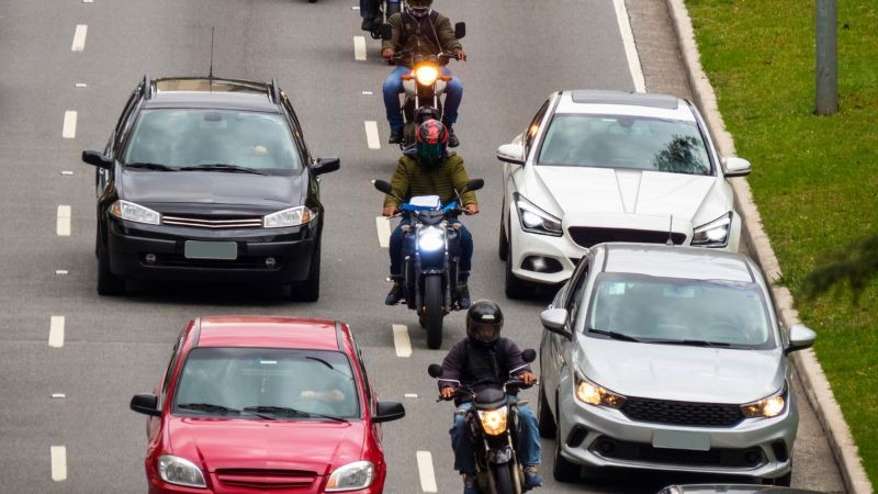carros e motos circulando em uma via de três faixas de mesmo sentido durante o dia 