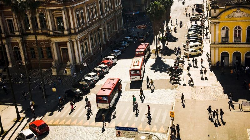 cruzamento de trânsito com microônibus vermelhos, veículos estacionados e pedestres atravessando a faixa de segurança