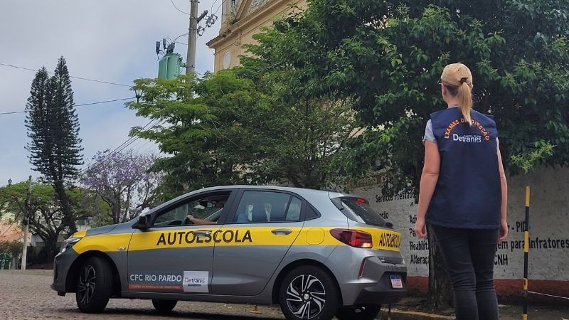 Foto horizontal colorida mostra um exame prático de direção. Carro de autoscola realiza manobra da baliza enquanto a examinadora de colete do DetranRS observa