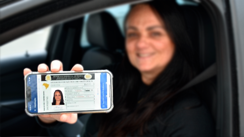 Foto horizontal colorida mostra condutora sorrindo pela janela do carro mostrando para  a câmera a tela do celular com a Carteira Nacional de Habilitação