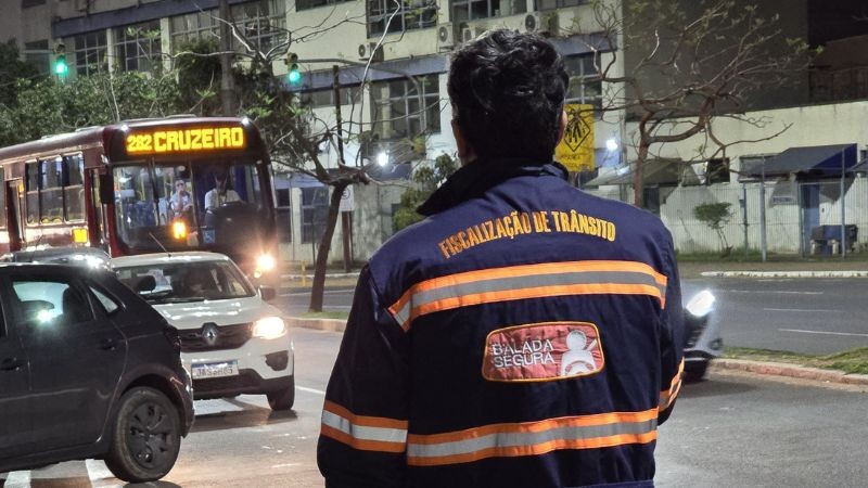 Foto horizontal colorida mostra agente de trânsito de costas com o colete da Balada Segura. Ao fundo, cena de trânsito noturno com três carros e um ônibus da linha Cruzeiro