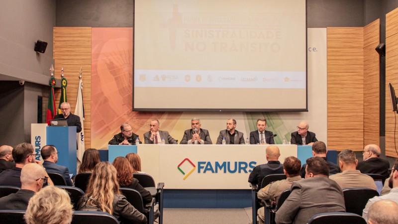 Foto horizontal colorida mostra mesa de evento com autoridades sentadas e a plateia  sentada de costas