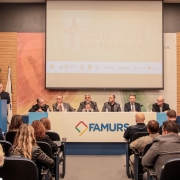 Foto horizontal colorida mostra mesa de evento com autoridades sentadas e a plateia  sentada de costas