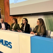 Foto horizontal colorida mostra três mulheres e um homem sentado em mesa de evento com o logo da Famurs 