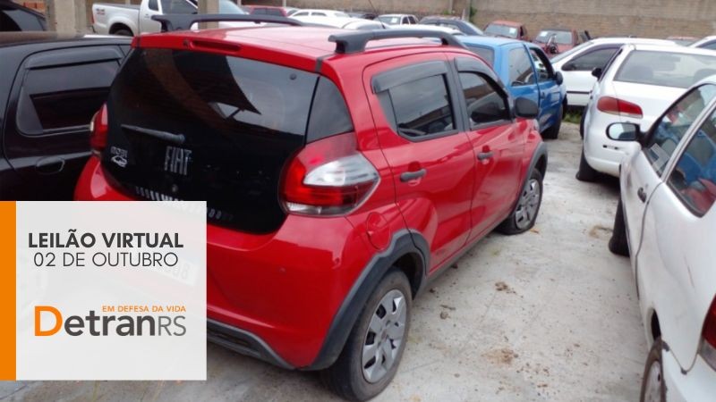 Fiat Mobi Vermelho com preto estacionado no centro entre outros veículos em um pátio de depósito e box cinza no canto inferior esquerdo onde está escrito leilão virtual 02 de outubro detranrs em defesa da vida, com pequena faixa lateral esquerda na vertical em laranja. 