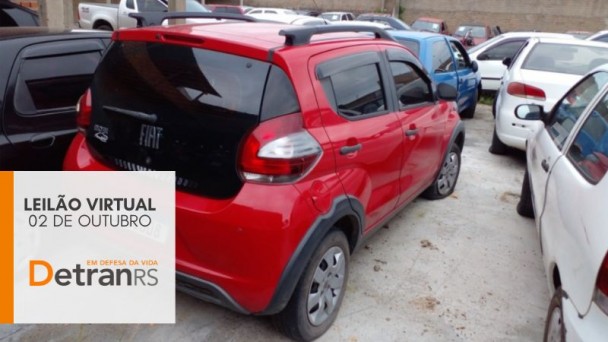 Fiat Mobi Vermelho com preto estacionado no centro entre outros veículos em um pátio de depósito e box cinza no canto inferior esquerdo onde está escrito leilão virtual 02 de outubro detranrs em defesa da vida, com pequena faixa lateral esquerda na vertical em laranja. 