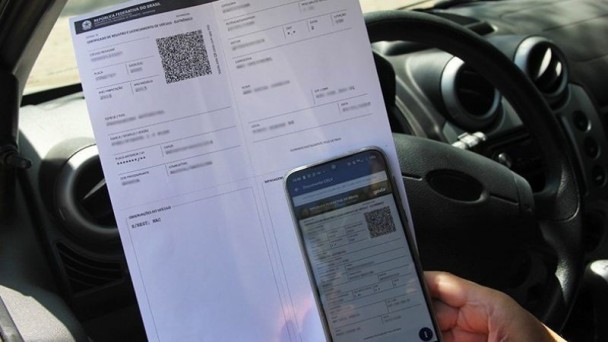 Celular com CRLV-e na tela sobre um crlv-e impresso em folha a4, todos em frente ao volante de um veículo