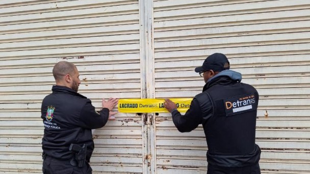 Dois homens de costas colocam lacre com logo do Detran na porta de estabelecimento fechado