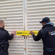 Dois homens de costas colocam lacre com logo do Detran na porta de estabelecimento fechado