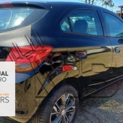 Veículo preto chevrolet ônix de lado/costas em primeiro plano, em estacionamento com outros veículos ao fundo. Box cinza com faixa laranja na lateral inferior esquerda onde está escrito leilão virtual 30 de outubro detranrs em defesa da vida.