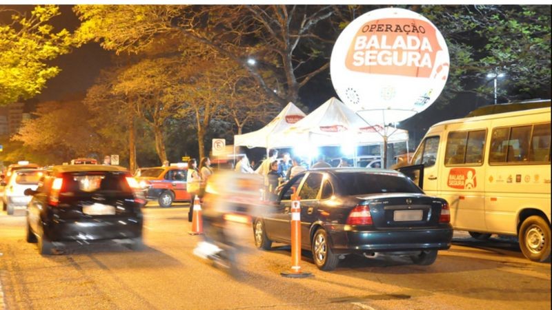 Veículos em duas faixas, passando por blitze da balada segura identificada por balão da operação, duas tendas e ônibus.