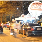 Veículos em duas faixas, passando por blitze da balada segura identificada por balão da operação, duas tendas e ônibus.