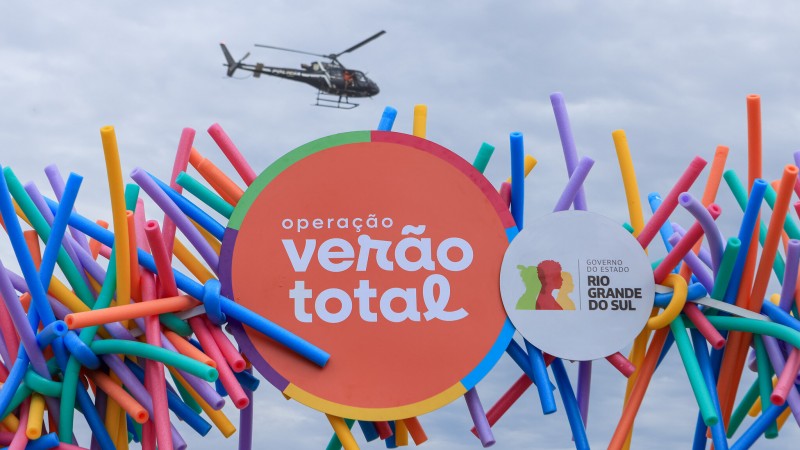 Foto horizontal colorida mostra painel com logo da Operação Verão Total. No fundo, um helicóptero voa em um céu nublado