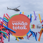 Foto horizontal colorida mostra painel com logo da Operação Verão Total. No fundo, um helicóptero voa em um céu nublado