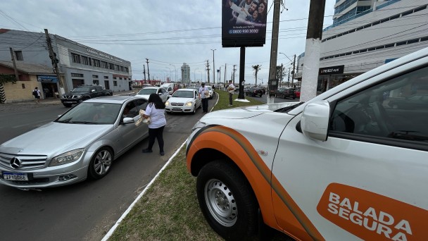 Foto horizontal colorida mostra o cenário de uma avenida movimentada com a vitura da Balada Segura parada no canteiro e agentes conversando com motoristas pela janela dos carros