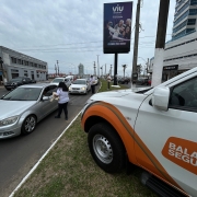 Foto horizontal colorida mostra o cenário de uma avenida movimentada com a vitura da Balada Segura parada no canteiro e agentes conversando com motoristas pela janela dos carros