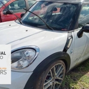 mini cooper branco quatro portas estacionado ao lado de peugeout vermelho em um depósito e box cinza onde está escrito leilão virtual 08 de janeiro