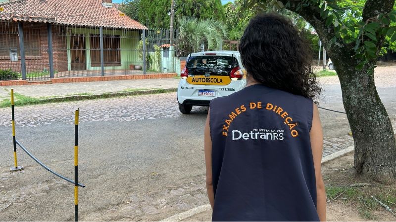 Foto horizontal colorida mostra mulher de costas com colete de examinadora do DetranRS, observando um carro de autoescola fazer baliza