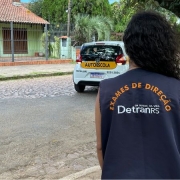 Foto horizontal colorida mostra mulher de costas com colete de examinadora do DetranRS, observando um carro de autoescola fazer baliza