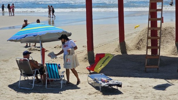 Foto horizontal colorida mostra um ambiente de praia com o mar e pessoas caminhando ao fundo. Em primeiro plano, uma mulher com a camiseta da Balada Segura entrega material edicativo para homem de boné sentado em uma cedeira de praia embaixo de um guarda sol ao lado da guarita do salva-vidas