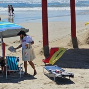 Foto horizontal colorida mostra um ambiente de praia com o mar e pessoas caminhando ao fundo. Em primeiro plano, uma mulher com a camiseta da Balada Segura entrega material edicativo para homem de boné sentado em uma cedeira de praia embaixo de um guarda sol ao lado da guarita do salva-vidas