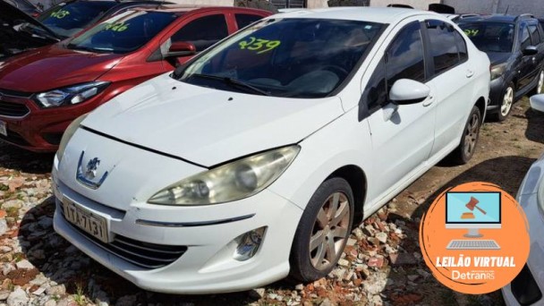 Foto horizontal colorida mostra carro branco estacionado em um páio com outros carros, todos com numeração no para-brisa.