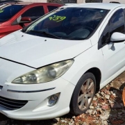 Foto horizontal colorida mostra carro branco estacionado em um páio com outros carros, todos com numeração no para-brisa.