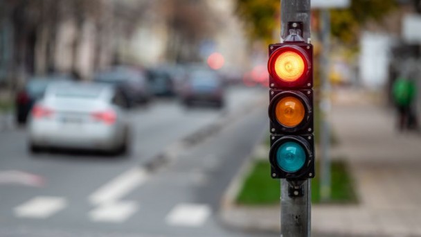 Foto horizontal colorida mostra semáforo no vermelho com ambiente de trânsito desfocado ao fundo