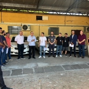 Foto horizontal colorida mostra grupo de pessoas em frente a fileira de carros doados. Homem de azul discursa e público olha pra ele.