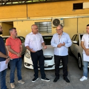 Foto horizontal colorida mostra cinco homens em frente a carros brancos estacionados. Homem de camisa rosa fala gesticulando.