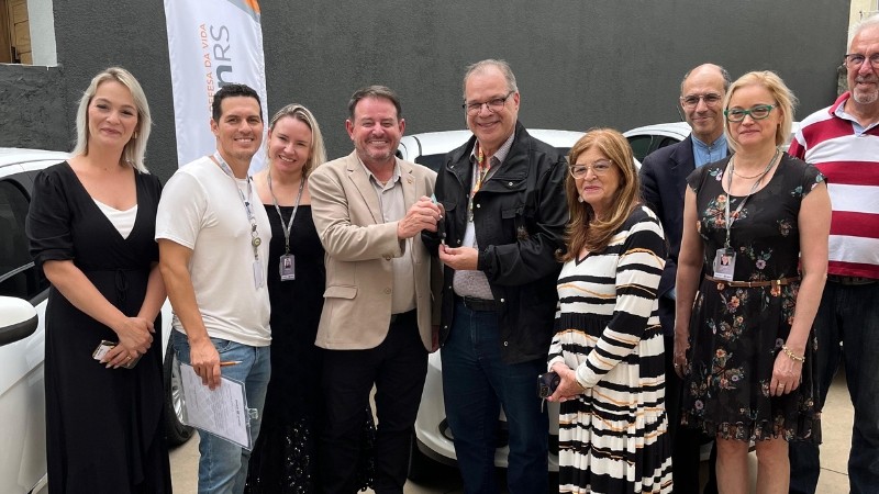 Foto horizontal colorida mostra grupo de pessoas posando para a fonto em frente a veículos brancos estacionados. No Centro um homem de casaco escuro entrega as chaves para homem de terno claro. Ao fundo, um wind banner do DetranRS.