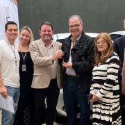 Foto horizontal colorida mostra grupo de pessoas posando para a fonto em frente a veículos brancos estacionados. No Centro um homem de casaco escuro entrega as chaves para homem de terno claro. Ao fundo, um wind banner do DetranRS.